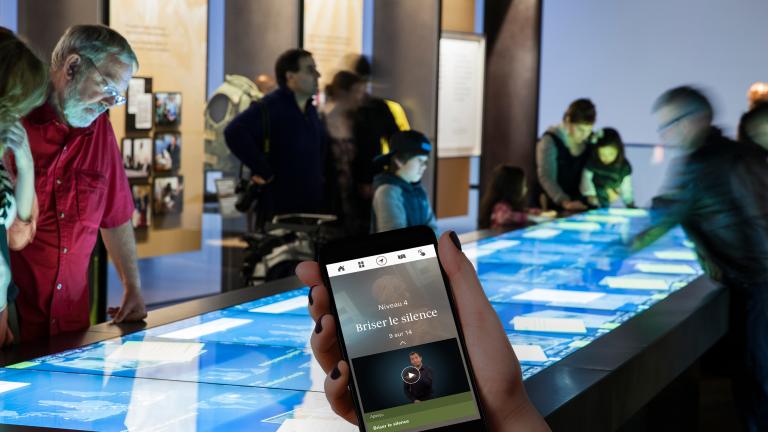 Une main tient un téléphone portable. Sur le téléphone se trouvent les mots « Niveau 4 » et « Briser le silence », et une personne parlant en langage des signes. En arrière-plan, des personnes regardent une grande table rectangulaire avec un écran tactile. Visibilité masquée.