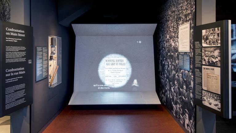Une exposition muséale comprenant des photos, du texte et un grand écran vidéo avec des sous-titres et une interprétation en langue des signes.