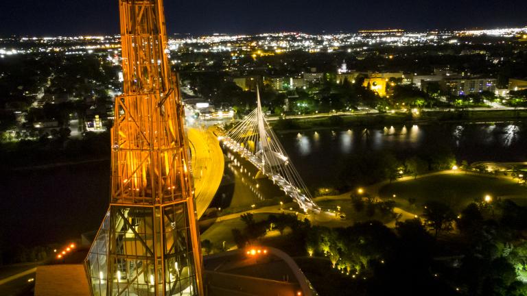 Une tour illuminée en orange vue d’en haut avec en arrière-plan un paysage urbain de nuit où on voit aussi une rivière et un pont.