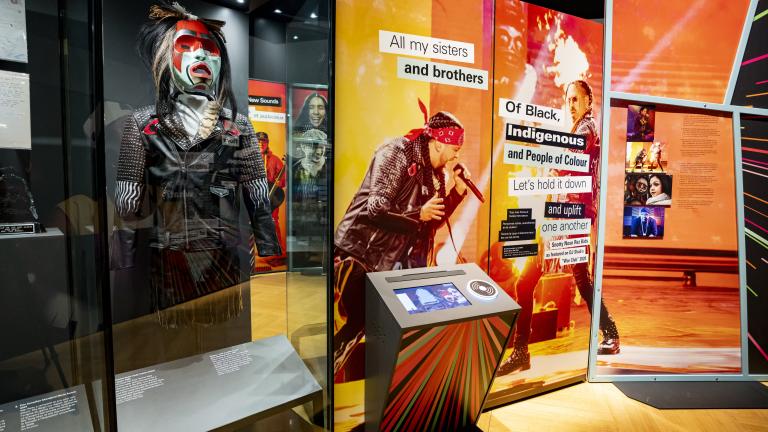 Une exposition muséale met en valeur des artistes autochtones de la musique. Elle contient une vitrine présentant un mannequin portant un masque et une veste, ainsi qu’un écran et un grand panneau graphique composé de textes et d’images sur fond orange.