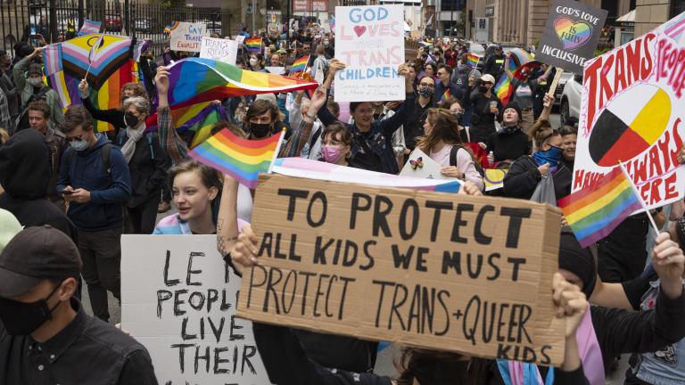 Un groupe de personnes se presse dans une rue de ville en brandissant des pancartes et des drapeaux arc-en-ciel. Sur la pancarte au premier plan, il est écrit en anglais : « Pour protéger tous les enfants, nous devons protéger les enfants transgenres et queer ».