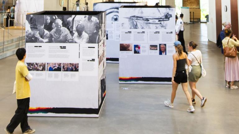 Trois grands cubes couverts d’images et de textes sont exposés dans une grande salle publique. Deux groupes de deux adultes s’approchent d’eux et consultent le contenu des cubes. Sur le côté, quatre personnes discutent.