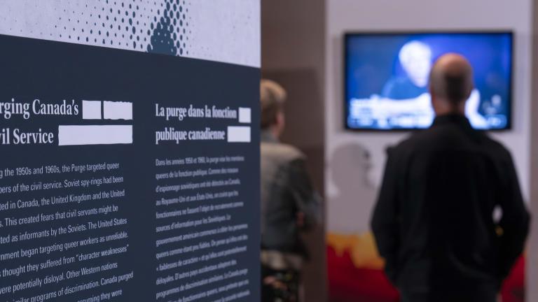 Un panneau d’exposition portant le texte « La purge dans la fonction publique canadienne » se trouve à gauche. Deux personnes à l’arrière-plan, à droite, regardent une vidéo.