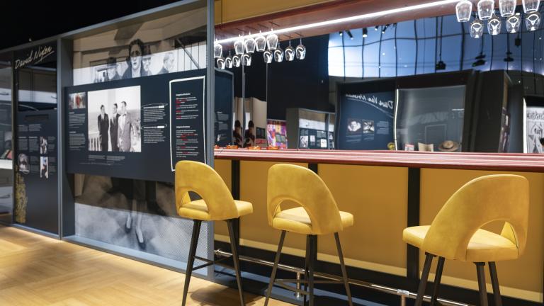 Une réplique d’un petit bar avec des tabourets hauts jaunes, un mur miroir derrière le bar et des verres à vin suspendus à des étagères en hauteur se trouve à côté d’un panneau d’exposition avec une grande image de personnes et du texte.