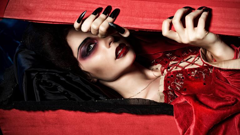Une vampire vêtue d’une robe rouge en velours regarde à travers une boîte en bois rouge qu’elle entrouvre avec ses longs ongles noirs. Visibilité masquée.