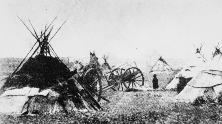 Une photo noir et blanc granuleuse de plusieurs tipis et d'une charrette en bois dans un champ. Visibilité masquée.