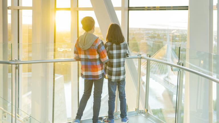 Deux enfants se tiennent dans une tour, dos à la caméra, et regardent vers l’horizon qui surplombe un paysage urbain. Visibilité masquée.