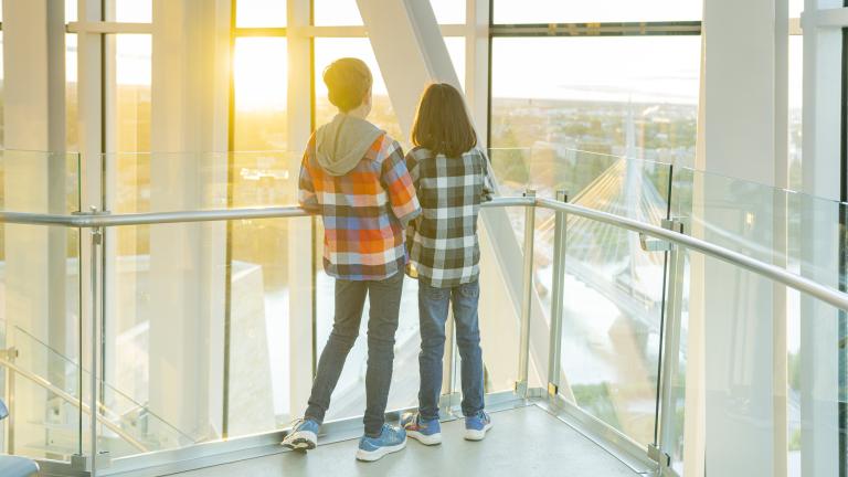Deux enfants se tiennent dans une tour, dos à la caméra, et regardent vers l’horizon qui surplombe un paysage urbain. Visibilité masquée.