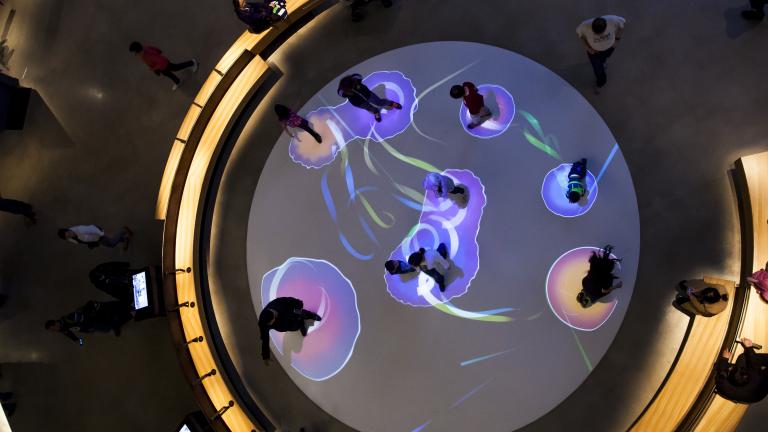 Une vue aérienne de personnes marchant sur un cercle blanc au sol. Chaque personne est entourée d'une bulle de lumière colorée projetée. Visibilité masquée.