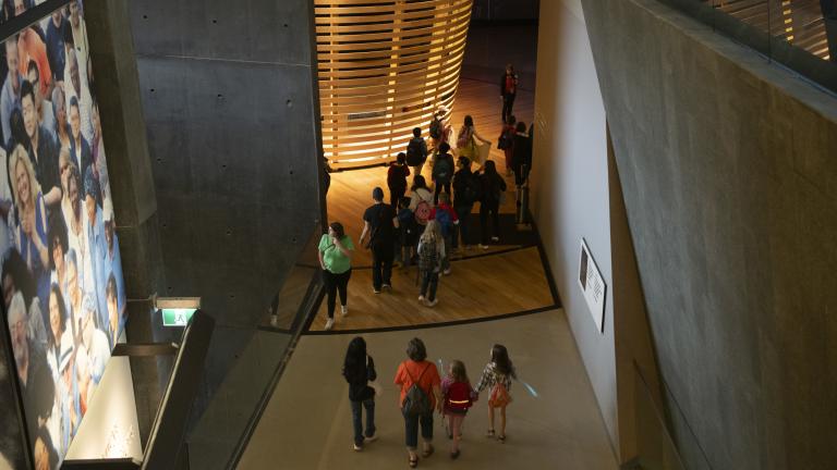 Des élèves se promènent dans les galeries du Musée. Sur le mur de gauche se trouve l’image d’une foule de personnes. Les murs sont en ciment. Visibilité masquée.