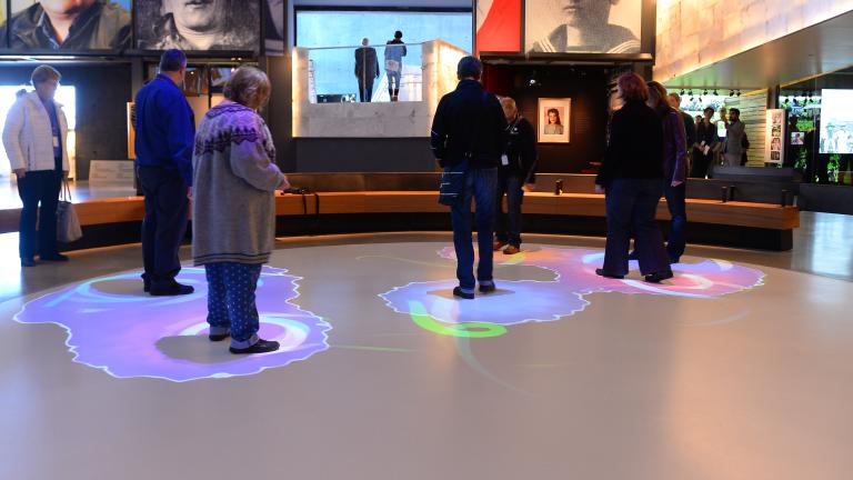 Un groupe de personnes se tient dans une partie d'un cercle blanc. Des lumières bleues et violettes sont projetées sur le sol autour de chaque personne. Visibilité masquée.