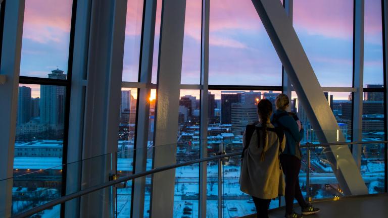 Deux personnes regardent par fenêtres allant du plancher au plafond un coucher de soleil sur un paysage urbain de Winnipeg. Visibilité masquée.