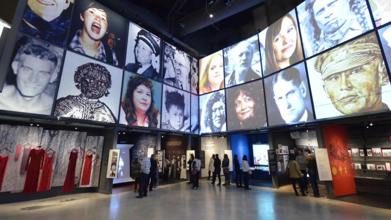 Les gens explorent une galerie du Musée avec deux rangées de portraits carrés au-dessus d'une série d'alcôves, contenant des photographies, des vidéos et du texte. L'alcôve de gauche contient des robes rouges suspendues. Visibilité masquée.