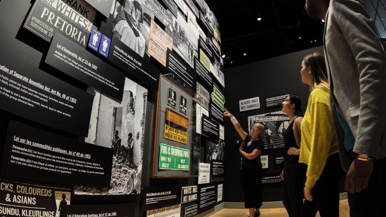 Quatre personnes d'âges et d'ethnies différents lisent les expositions du musée. Visibilité masquée.