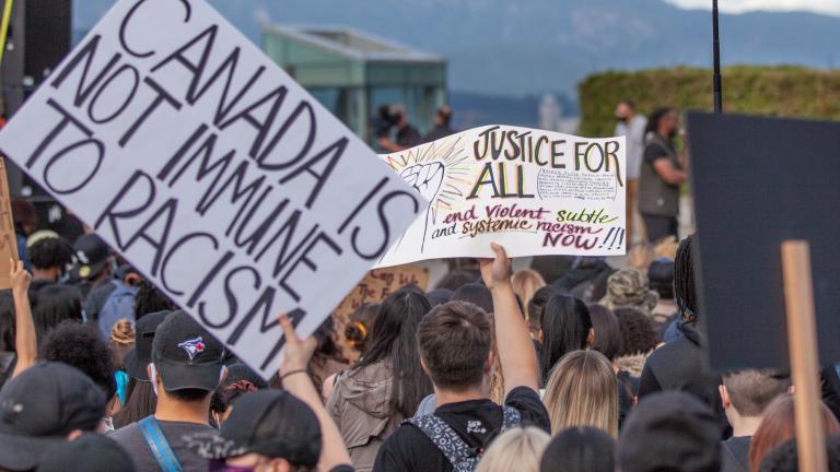 Une foule de personnes faisant dos à la caméra. Certains membres de la foule tiennent des pancartes audessus de leur tête qui disent, en anglais : « Je ne peux pas respirer #BLM », « Le Canada n'est pas à l'abri du racisme », et « Justice pour tous : mettez fin au racisme violent, subtil et systémique maintenant!!! ». Visibilité masquée.