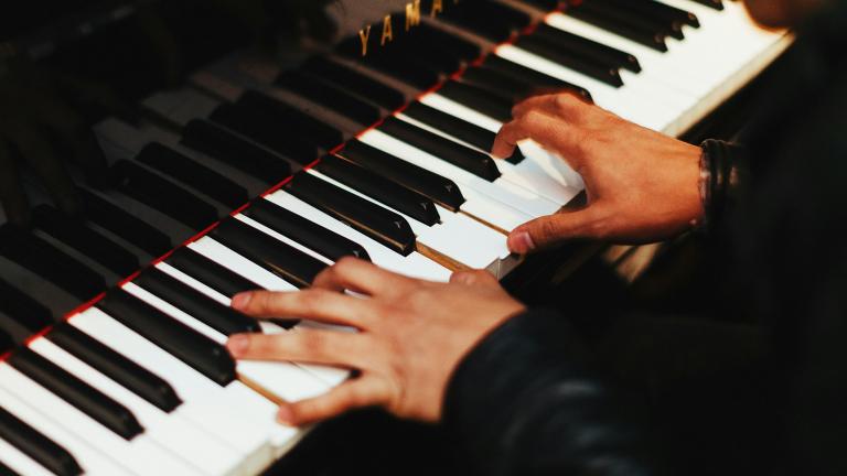 Deux mains jouent sur les touches d’un piano Yamaha d’un noir étincelant. La personne au piano, que l’on voit à peine, porte un haut noir à manches longues. Visibilité masquée.