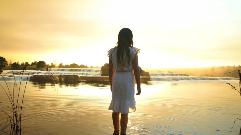 Une jeune fille aux longs cheveux noirs, vêtue d'une robe blanche jusqu'aux genoux, regarde au loin, debout jusqu'aux chevilles dans un lac calme, le soleil couchant jetant une chaude lueur jaune sur l'eau. Visibilité masquée.
