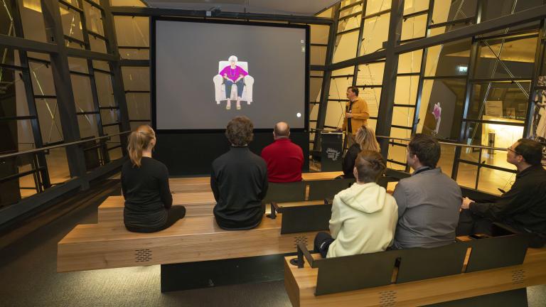 Un petit groupe de personnes assises sur des bancs en bois dans un théâtre fait face à un écran montrant une femme âgée assise dans un fauteuil. Un homme vêtu d’une chemise jaune se tient à droite de l’écran et tient un microphone. Visibilité masquée.