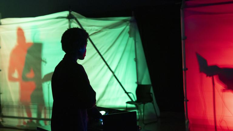 La silhouette d’une personne debout dans le noir devant des structures en tissu rouge et vert illuminées de l’intérieur et contenant des silhouettes ombragées. Visibilité masquée.