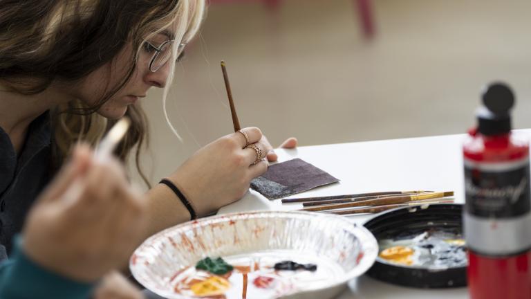 A young woman sitting at a table paints a design on a tile while a tin tray with various paints sits in front of her. An open bottle of red paint sits on the table. Visibilité masquée.