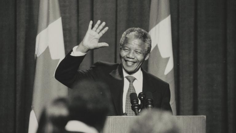 Une photographie en noir et blanc de Nelson Mandela debout derrière un podium. Il sourit et fait signe de la main. Visibilité masquée.