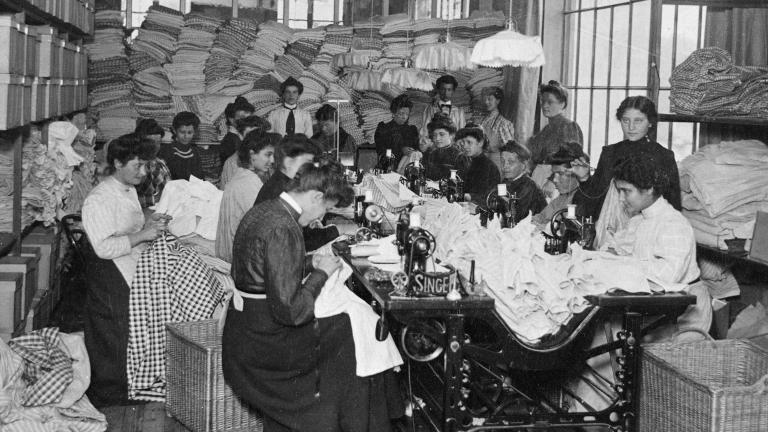 Un grand groupe de femmes assises en train de coudre. D’autres sont debout derrière elles dans une salle remplie de tissus. Visibilité masquée.