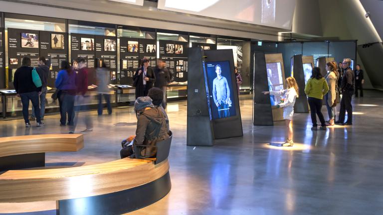 Des gens explorent une galerie contenant des vidéos interactives verticales au centre de la salle et des présentoirs avec texte et images qui longent le mur arrière.