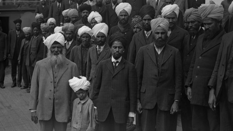 Une image en noir et blanc d’un grand groupe d’hommes sikh et d’un garçon debout sur le pont d’un navire. Ils font tous face au photographe et ne sourient pas.