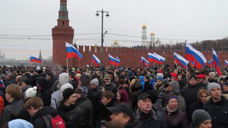 Une foule marche; plusieurs personnes portent le drapeau aux rayures rouge, blanche et bleue de la Russie. Derrière, on voit un mur de béton rouge qui se termine en flèche.