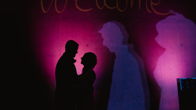 Les silhouettes d’un homme et d’une femme qui se font face et se tiennent devant un mur sombre éclairé de taches de lumière violette. Visibilité masquée.