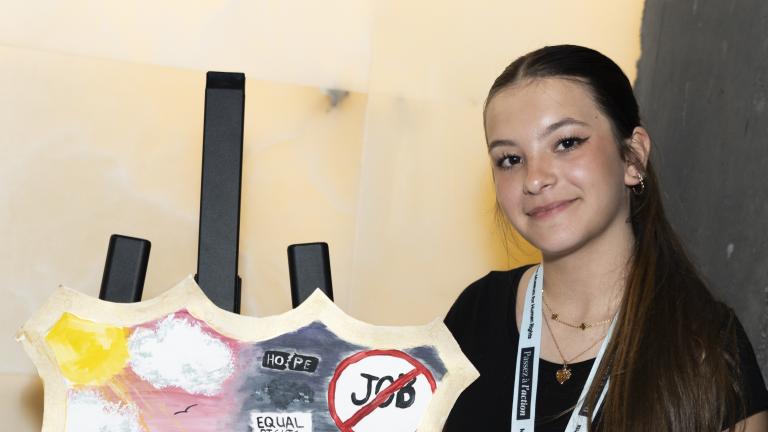 Une jeune femme se tient à côté d’un bouclier peint représentant d’un côté un ciel ensoleillé et un bateau flottant sur l’eau, et de l’autre un ciel sombre et un bateau en train de couler.