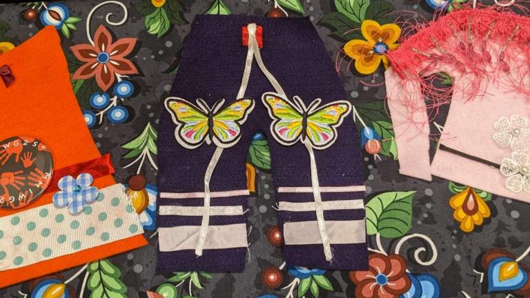 Des pièces de vêtements artisanaux de style autochtone sont posées sur un morceau de papier artisanal orné de fleurs et de baies. Les vêtements comprennent une chemise orange, un pantalon et une tunique rose décorée de tissus, d'appliques et de rubans colorés. La tunique comprend un châle à plumes sur les épaules. Visibilité masquée.