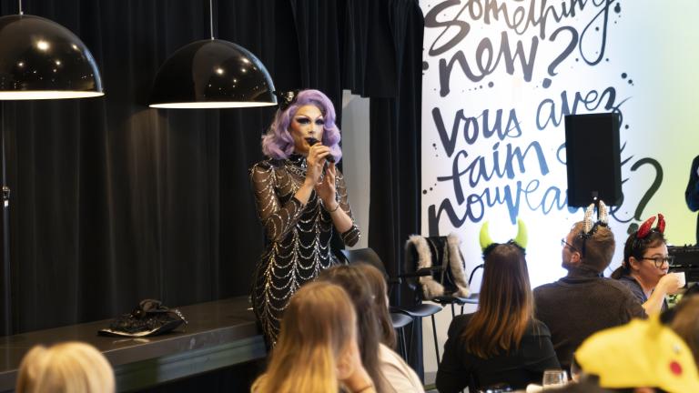 Une artiste de drag aux cheveux violets et à la robe argentée et noire s'exprime devant un groupe qui brunch. Visibilité masquée.