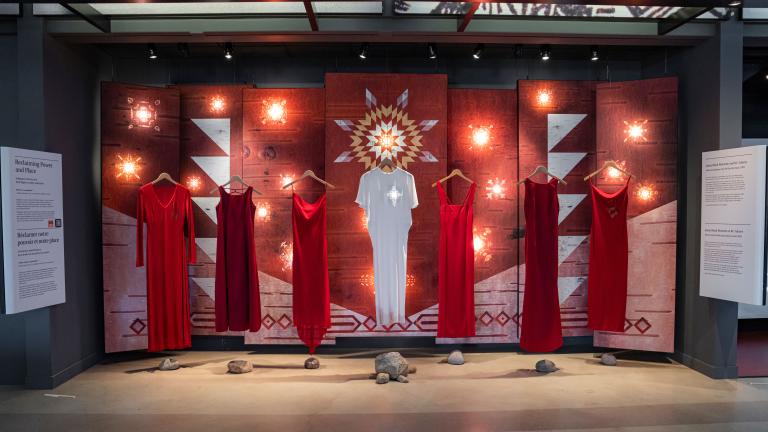 Six robes rouges et une robe blanche au milieu sont suspendues à des cintres devant une toile de fond. Une étoile dorée est projetée sur le cœur de la robe blanche. La toile de fond est composée de panneaux de bouleau peints en rouge, avec un soleil blanc et jaune au milieu et des formes géométriques blanches sur les derniers panneaux. Visibilité masquée.