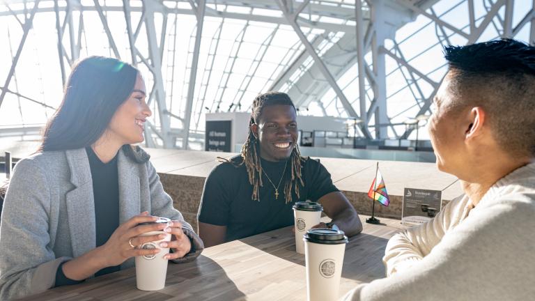 Trois personnes sont assises autour d'une table, souriantes, chacune avec une tasse à café en papier devant elle. Le soleil brille derrière elles, à travers les vitres qui entourent le Musée canadien pour les droits de la personne. Visibilité masquée.