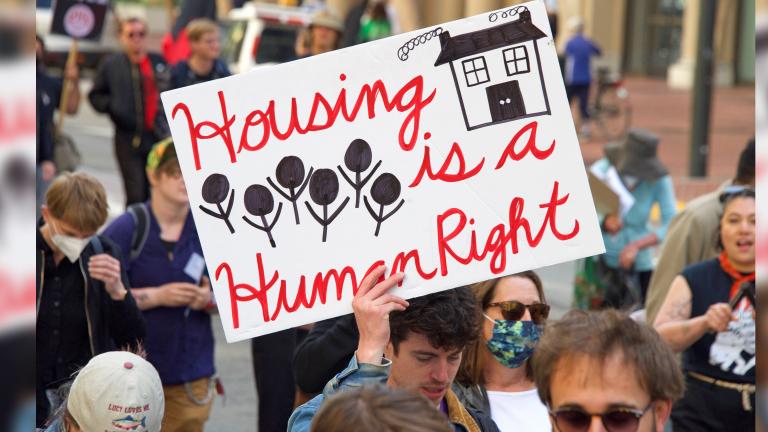 Un groupe de personnes marchant dans une rue. L’une d’entre elles tient une pancarte sur laquelle on peut lire : « Le logement est un droit de la personne. » Visibilité masquée.