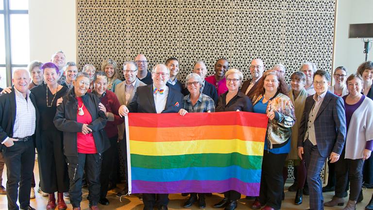  Vingt-huit membres du conseil d’ administration du Fonds Purge LGBT et du Comité consultatif du Monument national LGBTQ2+ posant ensemble et tenant un drapeau arc-en-ciel, lors de la première session de visualisation du Monument en 2019. Visibilité masquée.