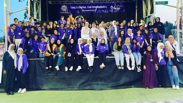 Des dizaines de personnes sont rassemblées sur une scène, la plupart souriantes et vêtues de violet. Beaucoup portent le hidjab. Une banderole est suspendue à l’arrière de la scène sur laquelle on peut lire, en anglais « Prenons l’initiative. Mettons fin à l’islamophobie. #OurLondonFamily ». Visibilité masquée.