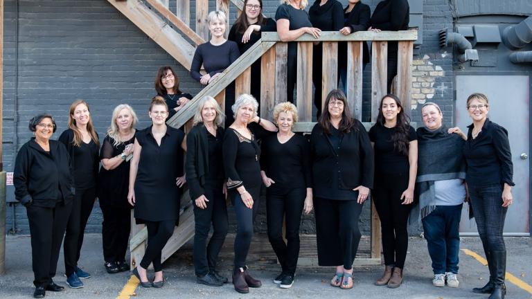 Les membres d’une chorale de femmes sont vêtus de noir et posent le long d’un escalier extérieur en bois. Visibilité masquée.