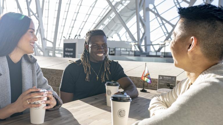Trois personnes sont assises à une table. Elles sourient toutes, un café devant elles. Un petit drapeau de la Fierté progressif est posé sur leur table et sur la table vide derrière elles. Le soleil brille à travers les fenêtres du Musée. Visibilité masquée.
