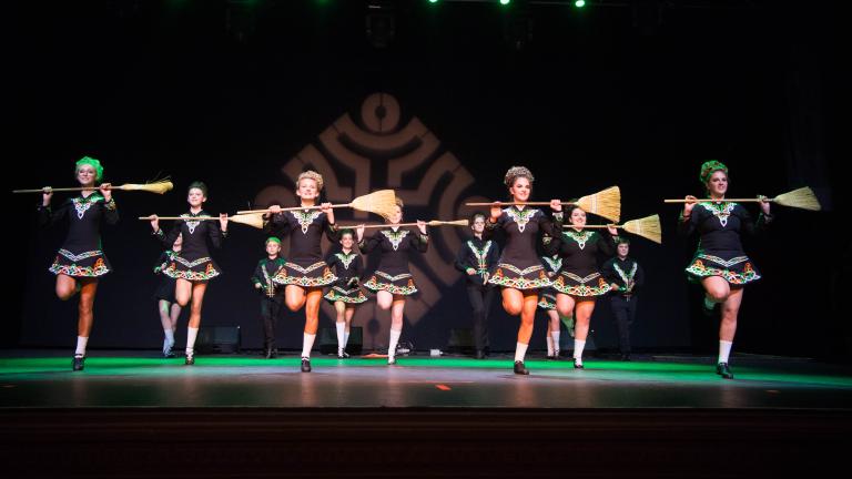 Treize danseuses irlandaises vêtues de courtes robes noires tiennent des balais sur leur poitrine. Visibilité masquée.