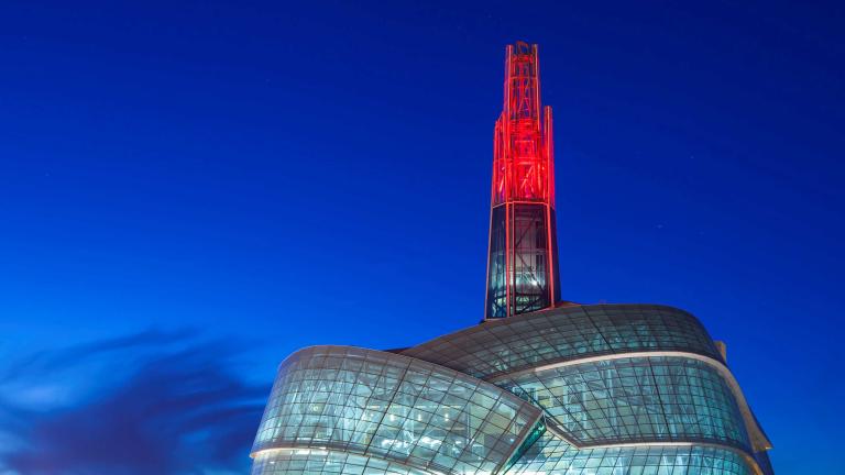 Un grand édifice avec une façade en verre incurvée et une tour en verre illuminée en rouge dans le crépuscule. Visibilité masquée.