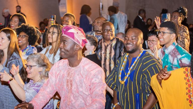 Un groupe de personnes de tous âges, dont beaucoup portent des vêtements africains traditionnels aux couleurs vives, dansent et sourient. Visibilité masquée.