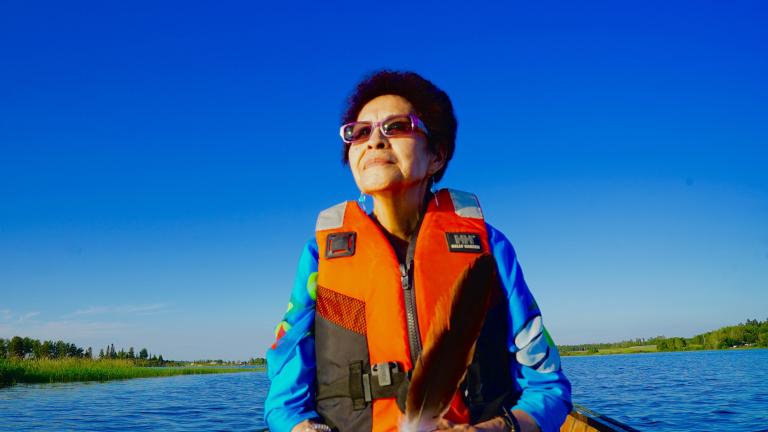 Une femme autochtone se trouve dans un canot sur un lac avec un gilet de sauvetage orange. Elle semble sereine. Visibilité masquée.