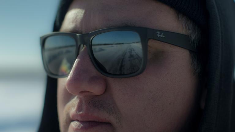 Un gros plan d'un homme portant des lunettes de soleil et un chapeau, qui regarde vers quelque chose hors champ. On voit le reflet d'une route dans ses lunettes. Visibilité masquée.
