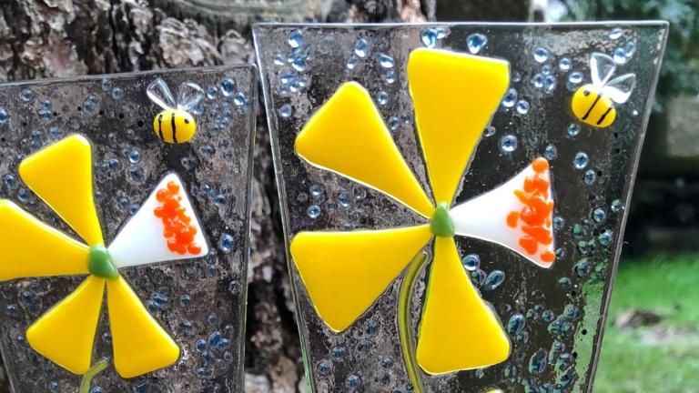 Deux décorations en verre fusionné représentant des fleurs jaunes sont placées dans un pot à fleurs à l’extérieur. Visibilité masquée.