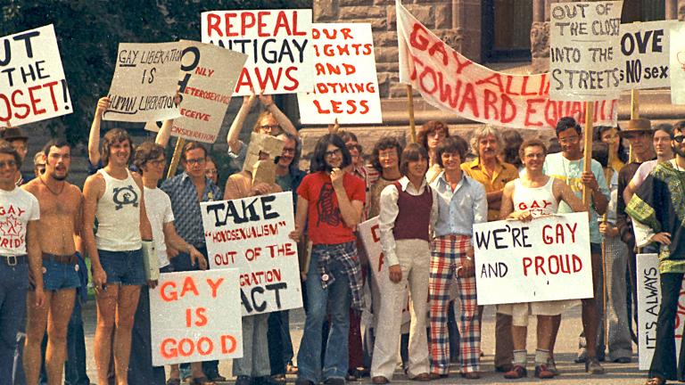 Un grand groupe de personnes vêtues de t-shirts et de pantalons évasés à la mode des années 1970 sont debout ensemble et tiennent des pancartes portant des messages en anglais tels que « sortis du placard », « c’est bien d’être gai » et « abrogez les lois anti-gaies ». Visibilité masquée.