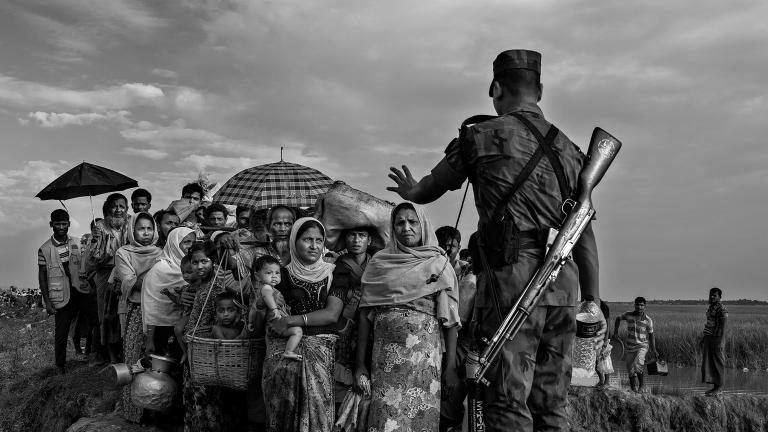 Un soldat avec un fusil en bandoulière sur le dos empêche un groupe de Rohingyas avant qu’ils ne traversent un pont de fortune. Sa main gauche est levée et fait un geste pour qu'ils s'arrêtent. Visibilité masquée.