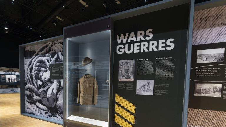 Une série de panneaux d’exposition présente des images historiques de militaires. Le mot « Guerres » figure en haut d’un des panneaux. Une vitrine située entre deux panneaux contient un uniforme militaire de la Première Guerre mondiale.