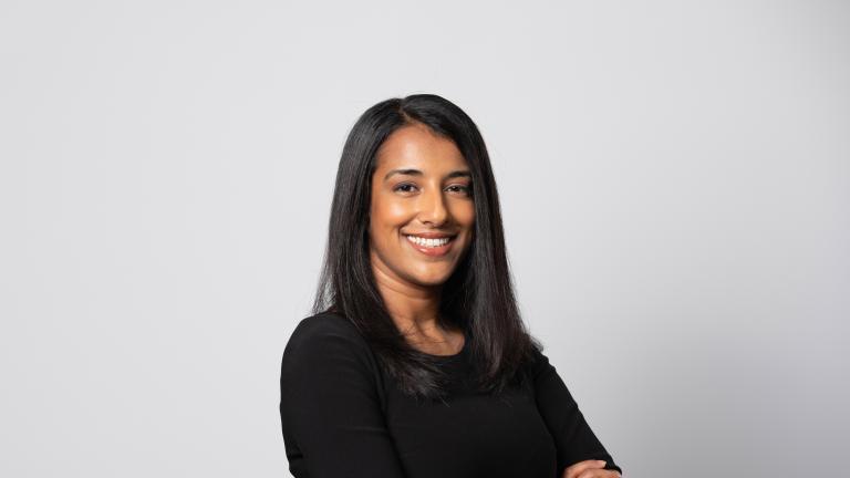 Le portrait d’une femme souriante aux bras croisés devant un arrière-plan blanc. Visibilité masquée.